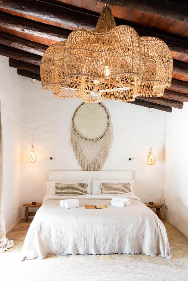 10 AUTHENTIC Finca Hotels in Ibiza 6 A cozy bedroom in the style of finca hotels in Ibiza, with a large bed, white bedding, woven pillows, pendant lights, a round macramé wall hanging, and a stunning woven chandelier beneath a wooden ceiling.