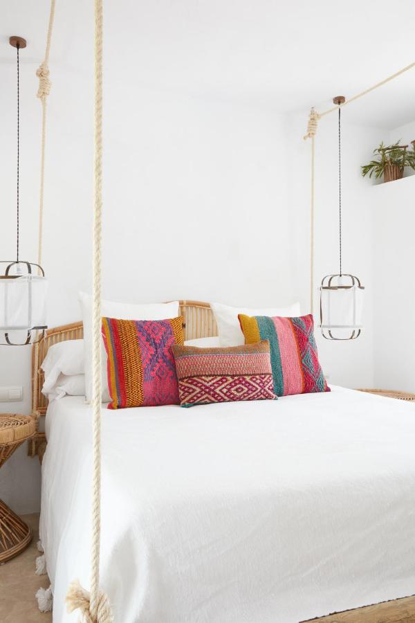 A bright, minimal bedroom inspired by boutique hotels in Formentera, with a bed featuring colorful patterned pillows, white bedding, and rattan details. The bed hangs from the ceiling with thick ropes and pendant lights on both sides.