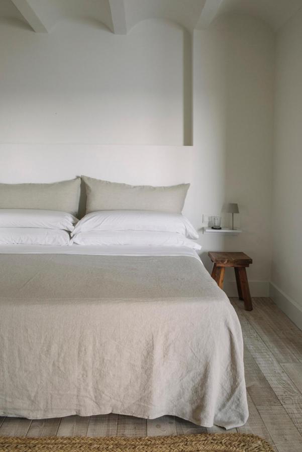 A neatly made bed with beige bedding and pillows is set against a white wall, evoking the calm style of boutique hotels in Costa Brava. A white shelf with a lamp, glass, book, and wooden stool sits beside the bed on a light wooden floor.