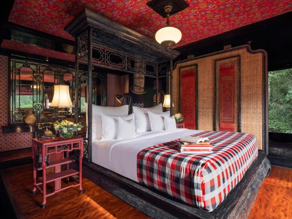 13 Most UNIQUE Places to Stay in Ubud 4 A luxurious bedroom with a large, dark wood canopy bed, white bedding, and a red plaid throw. This is one of the unique places to stay in Ubud, featuring ornate decor, patterned wallpaper, and large windows with views of lush greenery outside.