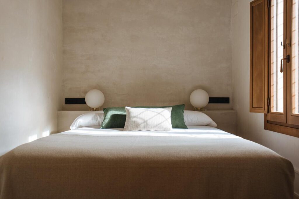 10 UNIQUE Hotels with Pool in Seville 6 A minimalist bedroom with a neatly made bed, neutral bedding, two green accent pillows, and round lamps on either side evokes the tranquil style found in top hotels with pool in Seville. Sunlight streams in from a wooden-framed window on the right.