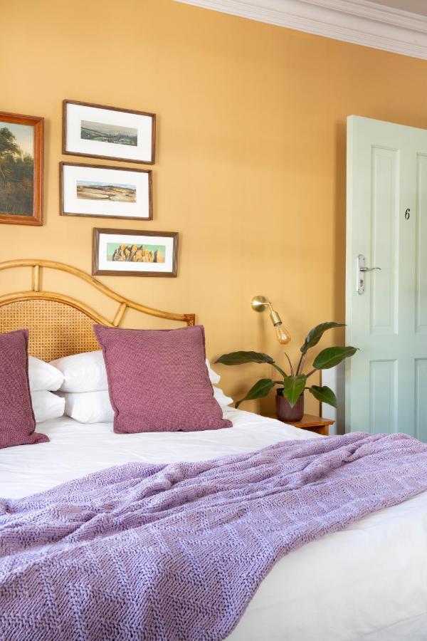 Unique Places to Stay in Cape Town 4 A cozy bedroom with a rattan headboard, white bedding, two purple pillows, and a lavender knitted throw—one of the unique places to stay in Cape Town. A plant and gold lamp sit on the bedside table beneath framed art on a mustard yellow wall.
