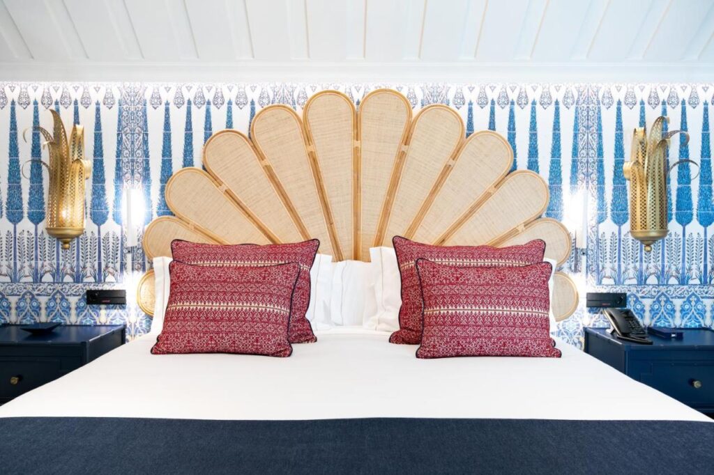 10 UNIQUE Hotels with Pool in Seville 21 A bed with a large, fan-shaped wicker headboard, three red decorative pillows, white bedding, and blue-and-white patterned wallpaper evokes the charming style of hotels with pool in Seville. Gold wall sconces are mounted on either side of the bed.