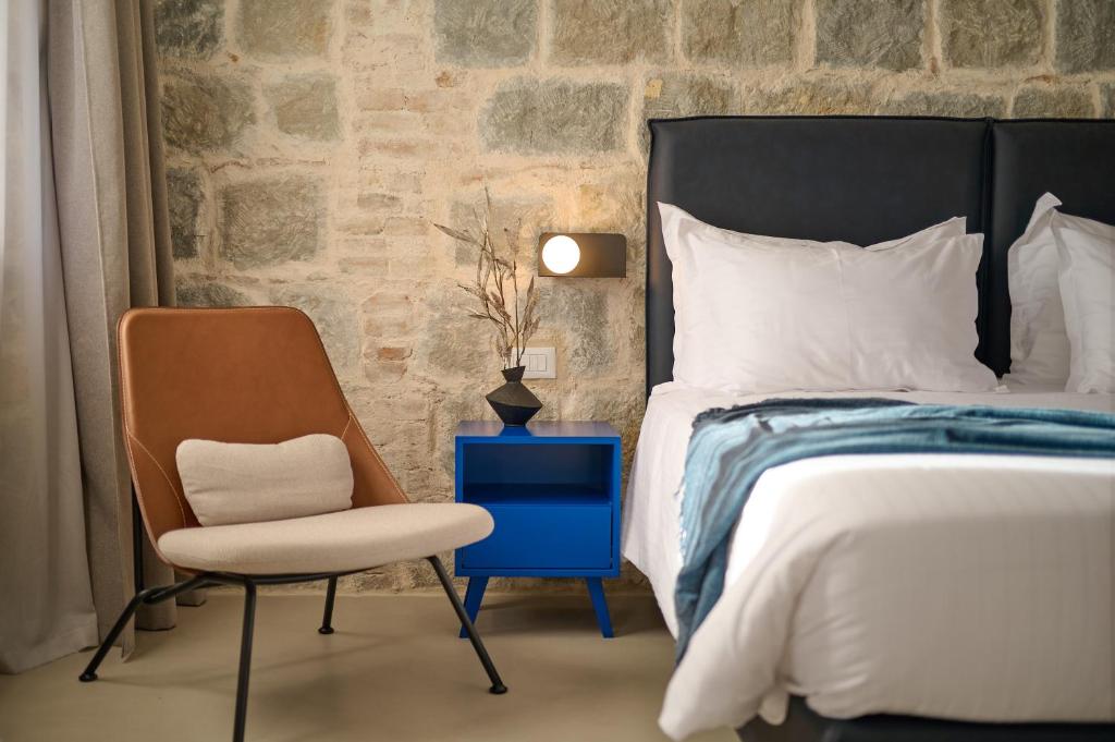 8 TOP Boutique Hotels in Split 18 A modern bedroom, perfect for those searching where to stay in Split, features a striking stone wall, a brown and beige chair, a blue nightstand with a vase of dried branches, a lit wall lamp, and a bed layered with white bedding and a blue throw blanket.