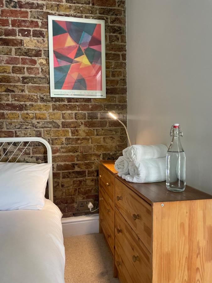 15 Best Boutique Hotels in London, UK 11 A bedside scene inspired by the best boutique hotels London offers, featuring a bed with white bedding, a wooden nightstand with neatly folded towels, a glass water bottle, a reading lamp, and colorful abstract art on an exposed brick wall.