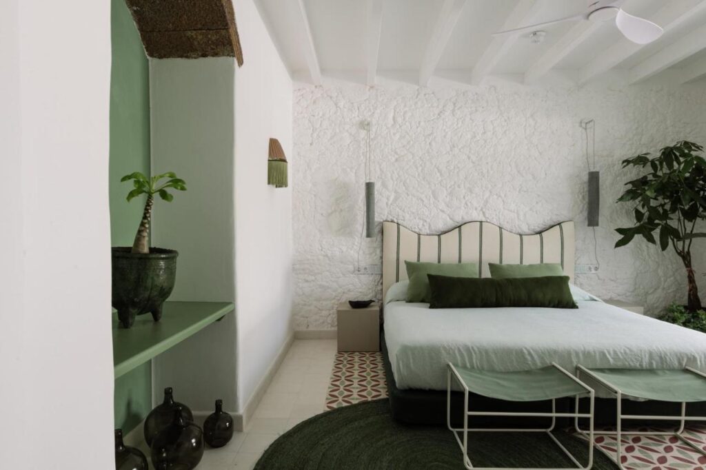 Unique Adults Only Hotels in Lanzarote 5 A modern bedroom inspired by Adults Only Hotels in Lanzarote, featuring a white stone wall, green and white decor, a potted plant, large bed with green pillows, round green rug, and bench. Natural light brightens the serene space.