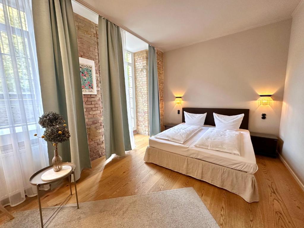 Boutique Hotels in Berlin Kreuzberg 28 A bright, modern bedroom inspired by boutique hotels in Berlin Kreuzberg, featuring a double bed, light wood flooring, brick accent walls, tall windows with green curtains, wall-mounted lamps, and a small round table with flowers.