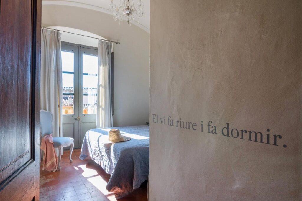 A sunlit bedroom in the style of boutique hotels in Costa Brava, with a blue armchair, a bed draped in a gray blanket and a straw hat on top. Light streams through tall windows with sheer curtains. The wall reads, “El vi fa riure i fa dormir.”.