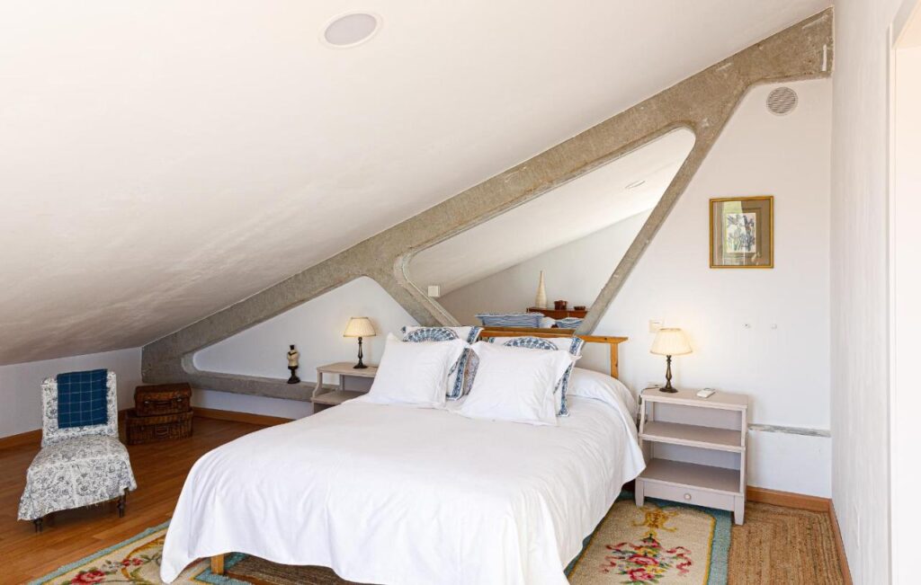 25 Weekend Trips from Madrid Max. 2h Away 30 A cozy attic bedroom with a slanted ceiling, double bed with white bedding—perfect for relaxing after weekend trips from Madrid. Two bedside tables with lamps, a patterned chair, wicker basket, and framed artwork complete the inviting space. Natural light brightens the room.