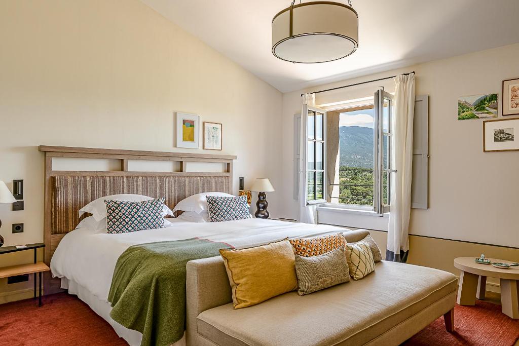 11 Unique Places to Stay in Provence 31 A cozy bedroom with a large bed, colorful pillows, a wooden headboard, and a bench at the foot. Sunlight streams through an open window, offering mountain views—one of the unique places to stay in Provence.