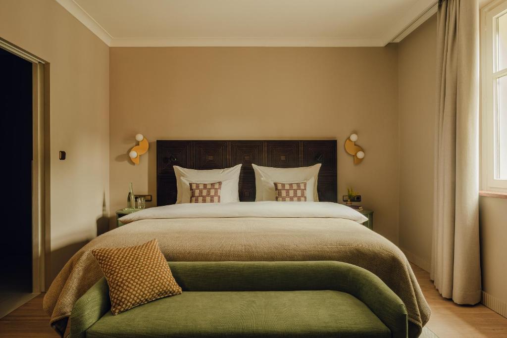 Unique Boutique Hotels in Salzburg 3 A cozy bedroom reminiscent of boutique hotels in Salzburg, with a large neatly made bed, patterned pillows, beige walls, bedside tables with lamps, and a green upholstered bench. Natural light streams in through curtained windows.