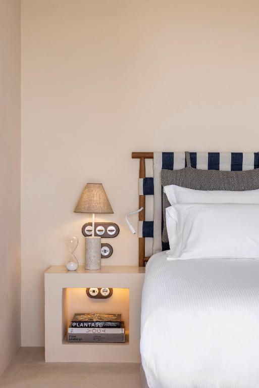Minimalist bedroom with a white bed, striped headboard, and built-in nightstand. Inspired by boutique hotels in Formentera, the neutral walls and bedding create a serene vibe. The nightstand holds a lamp, books, decor items, and has an open shelf underneath.