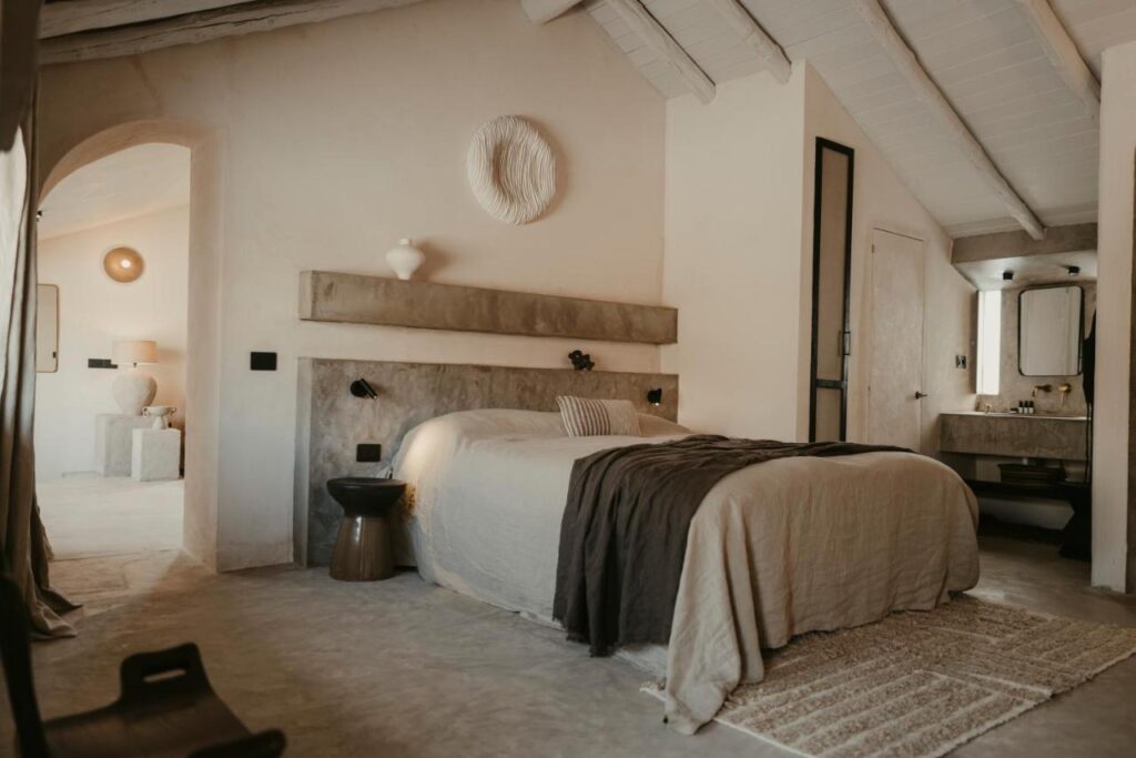 11 UNIQUE Boutique Hotels in Tenerife 5 A cozy, minimalist bedroom with beige walls and bedding, dark brown throw blankets, a modern headboard, and natural decor—reminiscent of the refined style found in boutique hotels in Tenerife. Warm, soft lighting illuminates a small vanity area and open doorway.