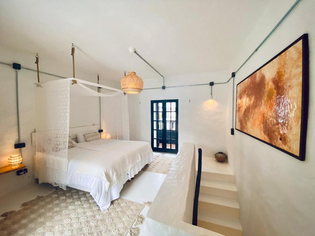 Unique Adults Only Hotels in Lanzarote 25 A bright, airy bedroom reminiscent of Adults Only Hotels in Lanzarote, with a white canopy bed, woven rug, wicker lamp, abstract art on the wall, and a black-framed glass door leading to a balcony. White stairs separate the bed area from the entrance.