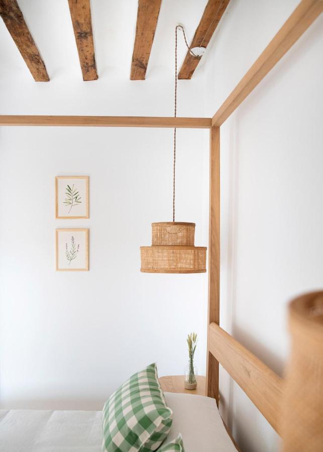 25 Weekend Trips from Madrid Max. 2h Away 73 Minimalist bedroom corner with exposed wooden ceiling beams, a rattan pendant lamp, two framed botanical prints—like scenes from weekend trips from Madrid—on a white wall, plus a bed with a green checkered pillow and small glass vase with flowers.