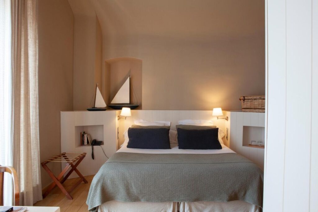 A cozy bedroom reminiscent of boutique hotels in Costa Brava, featuring a double bed with gray and white bedding, black pillows, built-in shelves displaying books and decor, model sailboats, bedside lamps, a wicker basket, and light brown curtains.