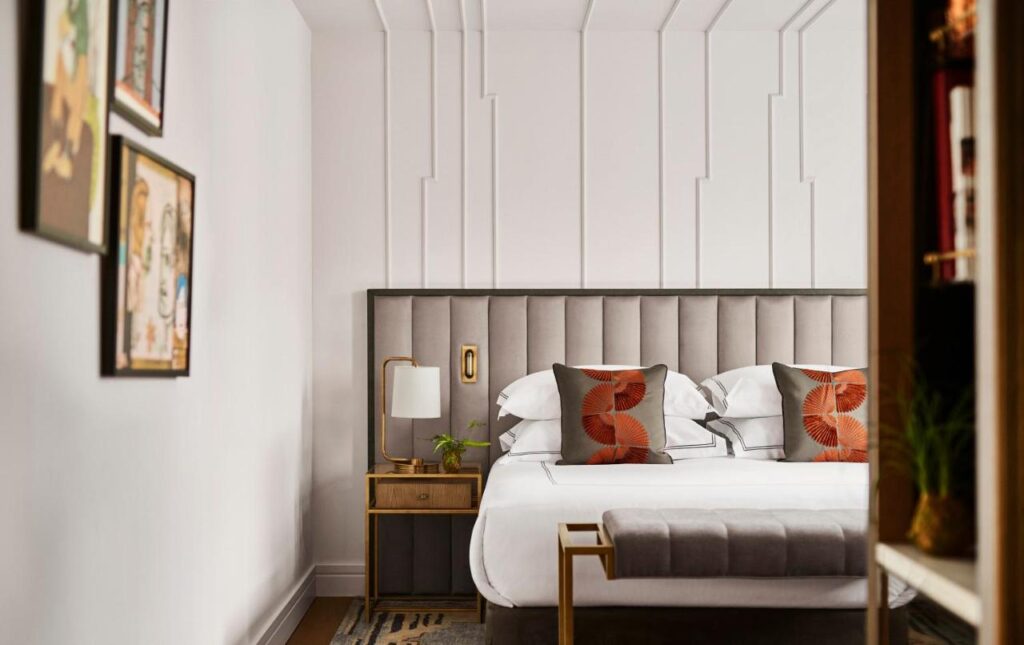 Where to Stay in Madrid: 14 Best Hotels 13 A modern bedroom—perfect inspiration for where to stay in Madrid first time—with a gray upholstered headboard, crisp white bedding, and bold orange pillows. A wooden nightstand with a lamp and plant sits beside the bed beneath framed art on the white wall.
