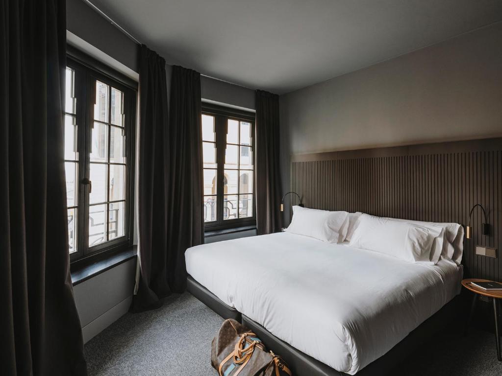 Minimalist hotel room with a large bed, white bedding, tall windows draped in black curtains, and a small wooden side table—exuding the modern, neutral-toned charm found in boutique hotels in Asturias.
