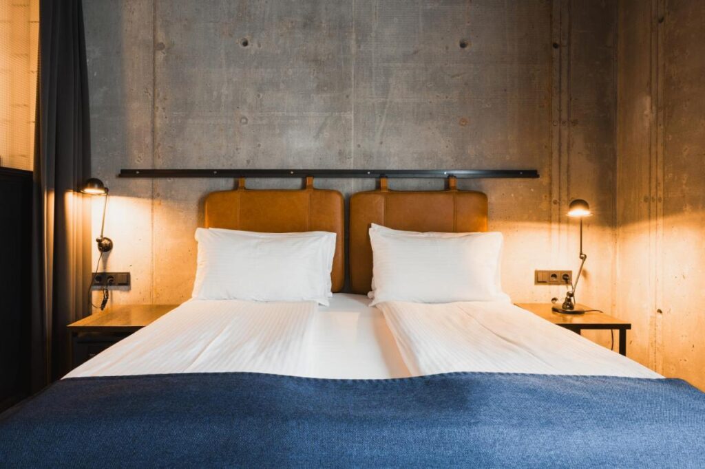 Where to Stay in Reykjavík: Top Guide 10 A modern hotel room—ideal for those searching where to stay in Reykjavik—features a double bed with white linens, brown headboards, a blue blanket, two bedside tables with lamps, and a concrete wall. The lighting is warm and inviting.
