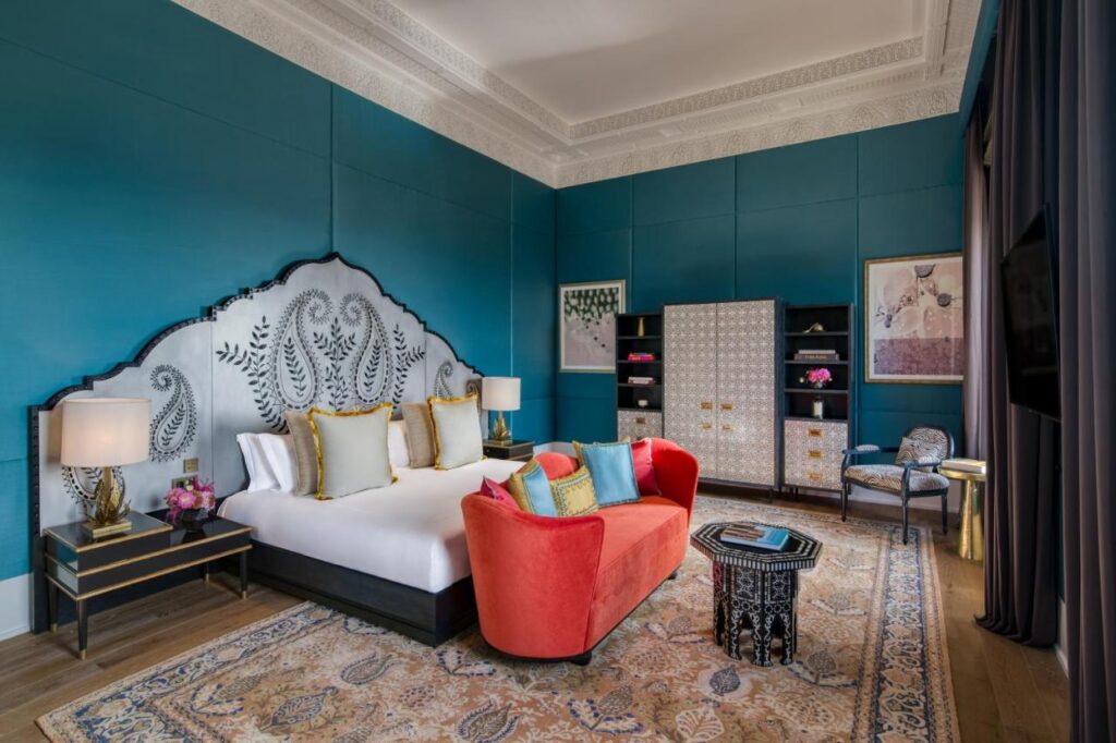 9 UNIQUE Riads in Tangier 7 Luxurious hotel room with a large bed, ornate headboard, teal walls, and red sofa—echoing the elegance of riads in Tangier—with decorative lamps, elegant storage cabinets, and framed artwork creating a stylish, inviting atmosphere.