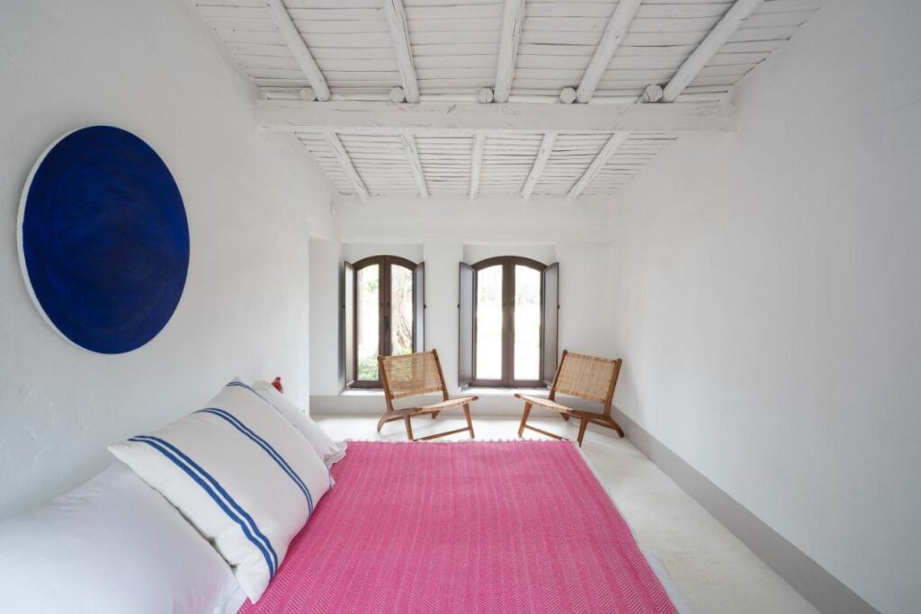 25 Weekend Trips from Madrid Max. 2h Away 10 A bright, minimalist room with white walls and ceiling, a bed with a pink cover and striped pillows, two wooden chairs by three arched windows—perfect for relaxing after weekend trips from Madrid—and a round blue artwork on the wall.