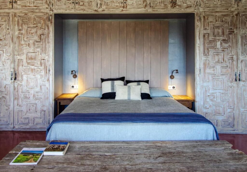 A cozy bedroom inspired by boutique hotels in Costa Brava, featuring a double bed with a wooden headboard, two bedside tables with wall-mounted reading lights, rustic wooden closet doors, and a bench with books at the foot of the bed.