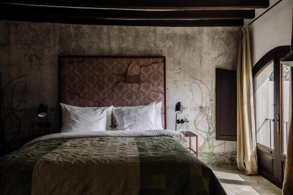 10 AUTHENTIC Finca Hotels in Ibiza 10 A rustic bedroom often found in finca hotels in Ibiza, with a double bed, white pillows, patterned headboard, textured walls, and a quilted green blanket. Sunlight pours in through an open window framed by thick curtains and wooden shutters.