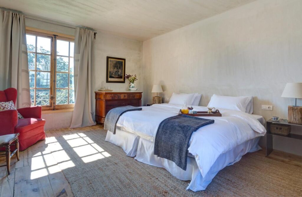 25 Weekend Trips from Madrid Max. 2h Away 51 Perfect for weekend trips from Madrid, this bright bedroom features two beds pushed together, white bedding, dark gray throws, a red armchair, wooden dresser, side tables with lamps, and a sunlit window with light curtains.