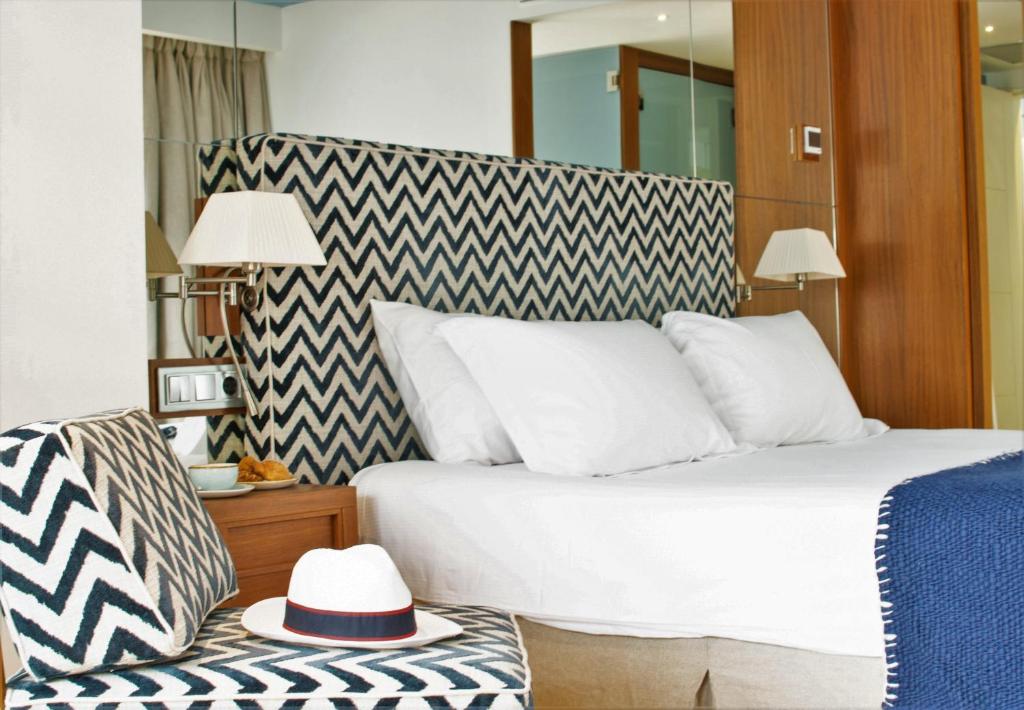 A modern hotel room at one of the boutique hotels in Formentera, featuring a bed with a zigzag-patterned headboard, white bedding, a matching chair with a white hat, wooden nightstand, wall lamps, and a blue blanket at the foot of the bed.