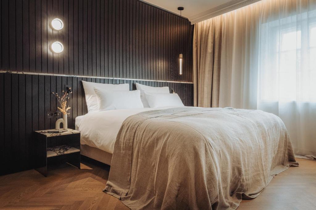 Unique Boutique Hotels in Salzburg 19 A cozy bedroom reminiscent of boutique hotels in Salzburg, featuring a large bed with white bedding, a beige blanket, dark wood panel walls, a nightstand with dried flowers, and soft natural light filtering through sheer curtains.