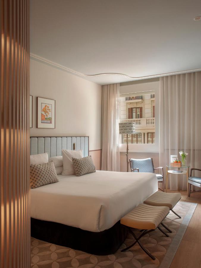 Top Barcelona Hotels with Rooftop Pool 23 A modern hotel room with a double bed, accent pillows, padded headboard, small bench, and armchairs by a round table near the window—perfect for relaxing after exploring Barcelona hotels with rooftop pools. Wall art hangs above the bed.
