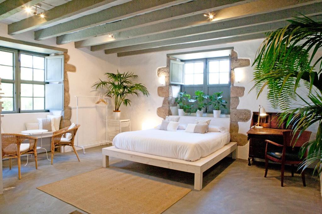 11 UNIQUE Boutique Hotels in Tenerife 19 A bright, airy bedroom inspired by boutique hotels in Tenerife, with exposed wooden beams, a large bed with white bedding, potted plants on the windowsill, wicker chairs at a small table, a wooden desk with a lamp, and sunlight streaming through shuttered windows.