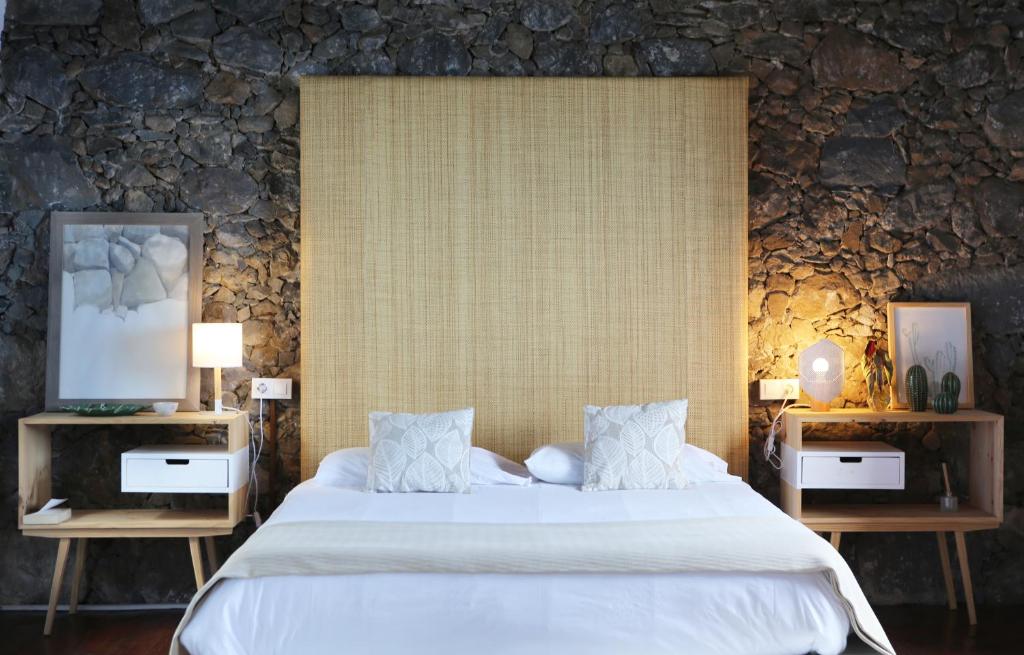 11 UNIQUE Boutique Hotels in Tenerife 38 A cozy bedroom inspired by boutique hotels in Tenerife, featuring a large beige headboard, white bed linens, and two wooden nightstands with lamps and décor, all set against a textured stone wall.