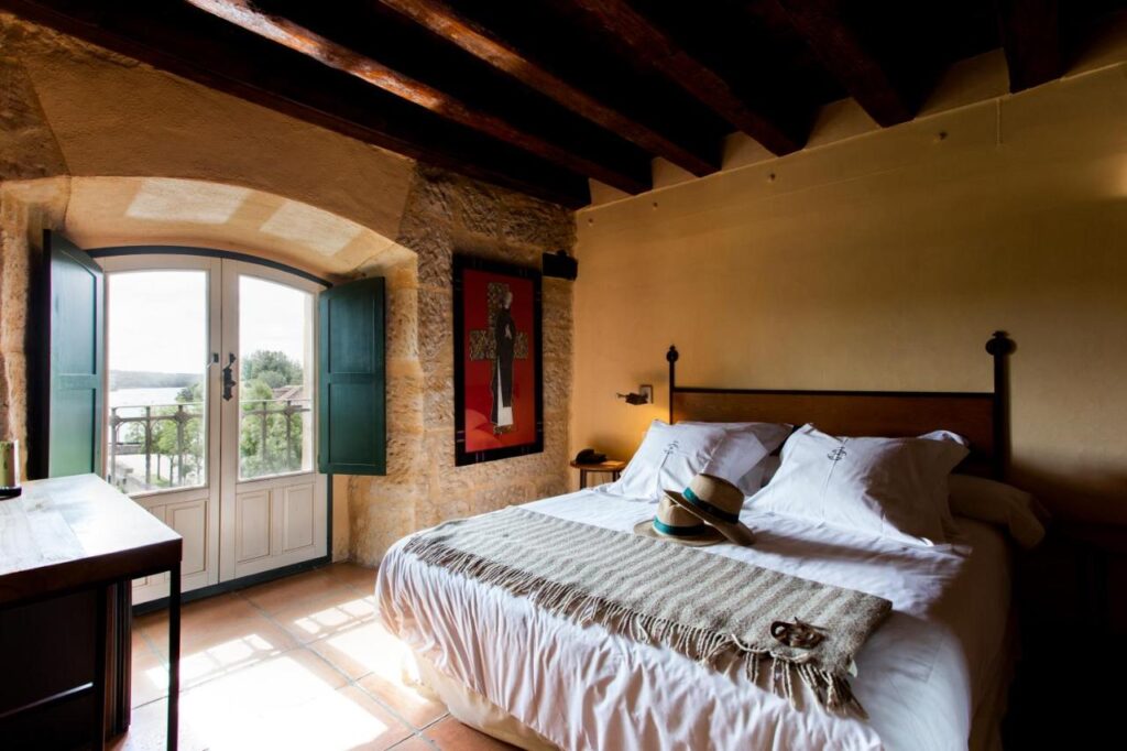25 Weekend Trips from Madrid Max. 2h Away 23 Cozy bedroom with rustic stone walls, beamed ceiling, a large bed with pillows and a throw blanket—perfect for weekend trips from Madrid. Two hats rest on the bed, with double doors opening to a scenic balcony view.