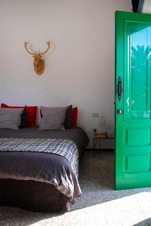 Unique Adults Only Hotels in Lanzarote 10 A cozy bedroom at one of the Adults Only Hotels in Lanzarote, with a double bed featuring gray and red pillows, a wooden deer head wall decoration above, and a bright green door open to sunlight. A small nightstand with a lamp sits beside the bed.