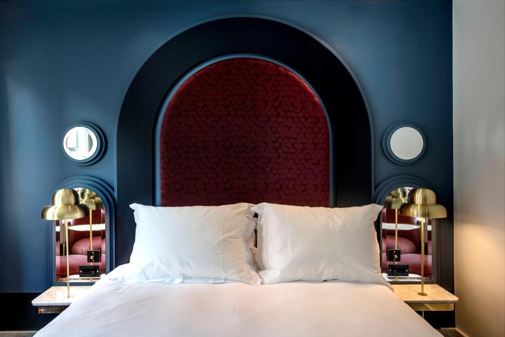 15 Best Boutique Hotels in London, UK 50 A neatly made bed with white pillows and sheets sits against a dark blue wall with a large arched red headboard—an inviting scene often found in boutique hotels in London. Two gold lamps and round mirrors sit on matching bedside tables.