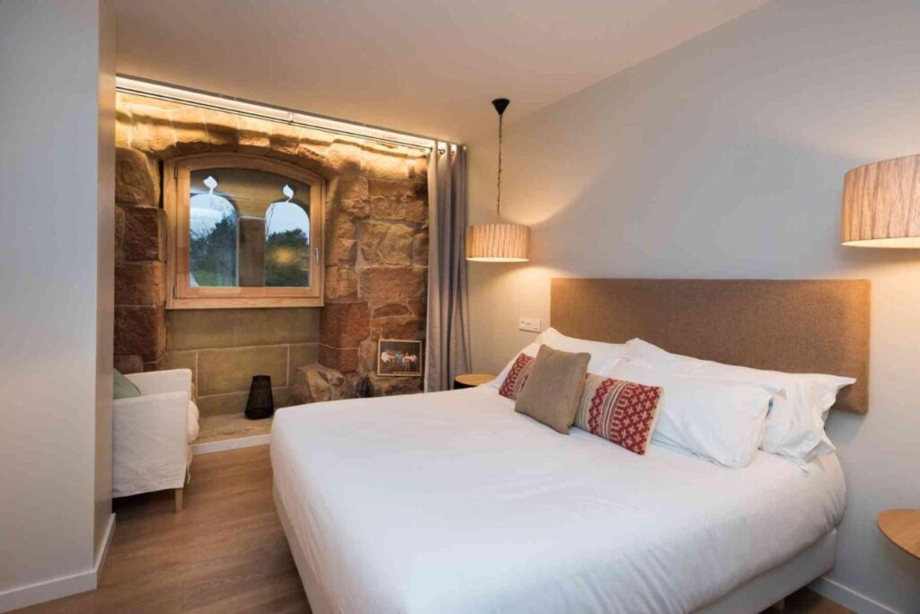 Best Boutique Hotels in San Sebastian 6 A cozy bedroom in the style of boutique hotels in San Sebastian, with a large bed, white linens, decorative pillows, and a stone wall featuring an arched window. Soft lighting and a beige armchair create a warm, inviting atmosphere.