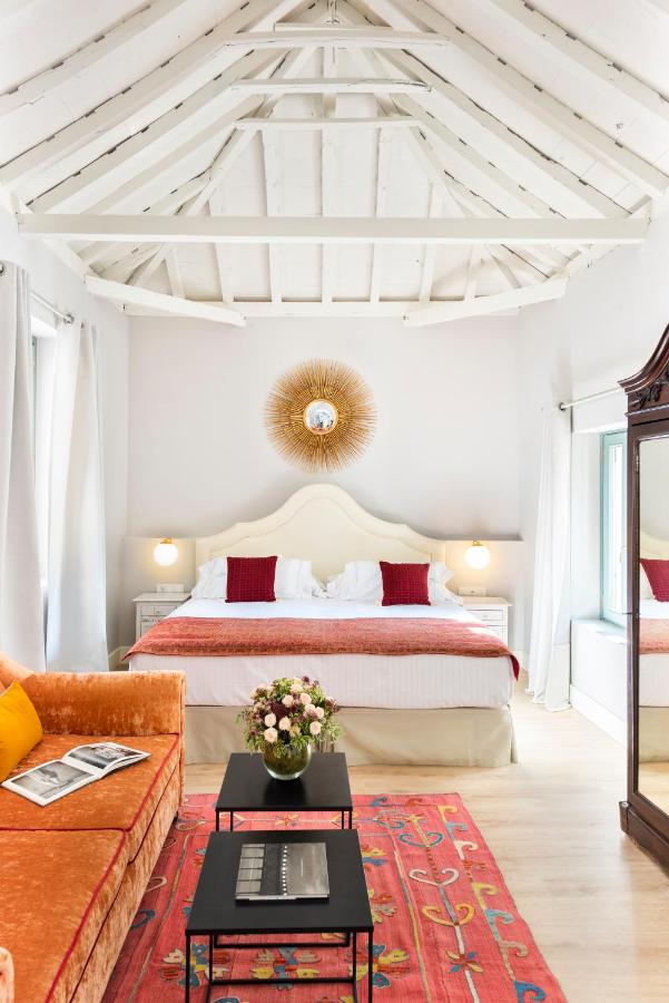 10 UNIQUE Hotels with Pool in Seville 32 A bright bedroom in the style of boutique hotels with pool in Seville, featuring a high white vaulted ceiling, a large bed with red pillows, a sunburst mirror above the headboard, an orange sofa, colorful rug, and fresh flowers on black coffee tables.