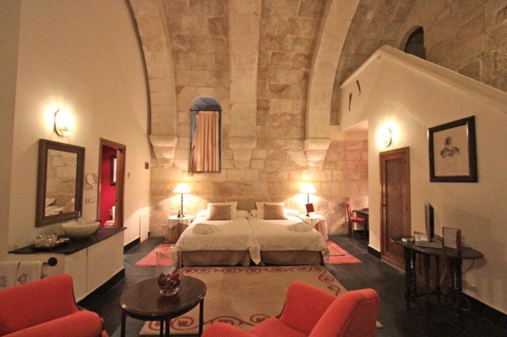 25 Weekend Trips from Madrid Max. 2h Away 27 A cozy, warmly lit bedroom with stone vaulted ceilings—perfect for weekend trips from Madrid—features a large bed with white linens, red armchairs, a rug, and elegant decor blending modern comforts and historical charm.