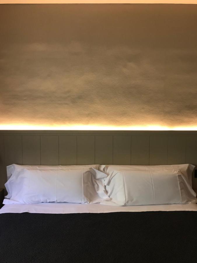 A neatly made bed with two white pillows and a white sheet, set against a wall with indirect warm lighting above the headboard—a calm, minimalistic scene inspired by boutique hotels in Costa Brava.