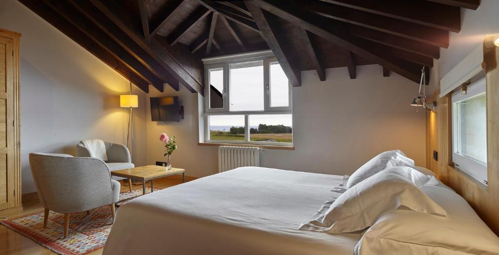 A cozy bedroom typical of boutique hotels in Asturias, featuring a large bed, two armchairs around a small wooden table, a wall-mounted TV, large windows with natural light, a dark wood-beamed ceiling, and a scenic outdoor view.