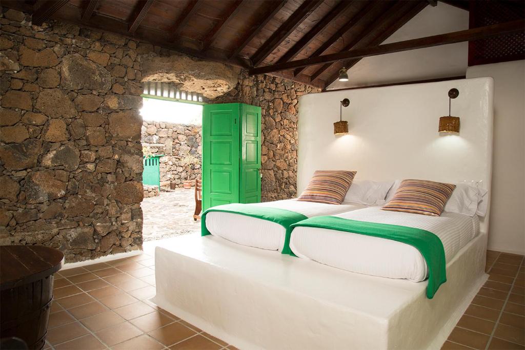 Unique Adults Only Hotels in Lanzarote 26 A rustic bedroom with stone walls, a wooden ceiling, two single beds with white linens and green throws—perfect for relaxing escapes at Adults Only Hotels in Lanzarote. Striped pillows, wall lamps, a green wardrobe, and an open door complete the tranquil scene.