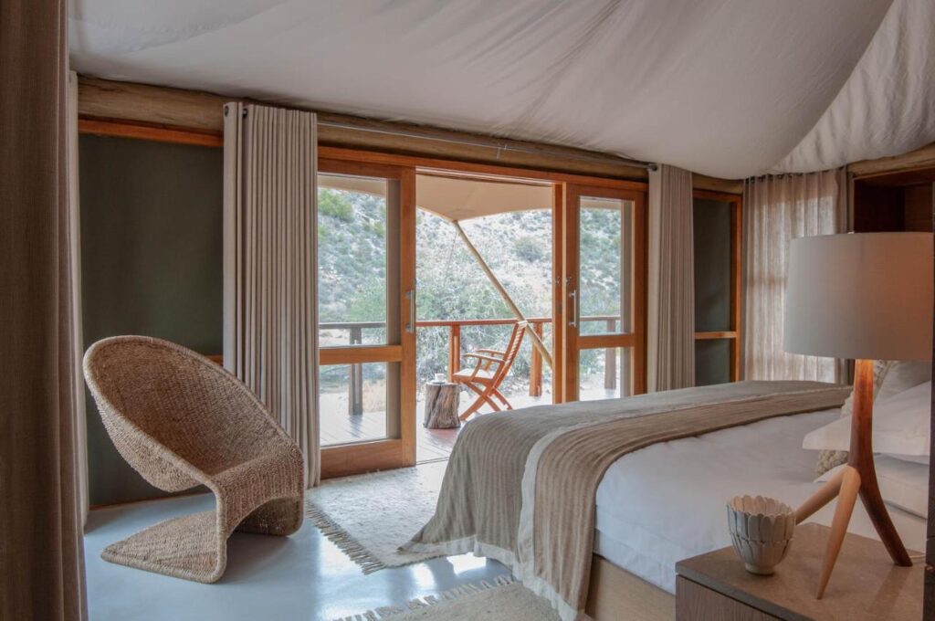 A cozy bedroom in one of the Game Lodges in Western Cape features a wicker chair, a double bed with beige linens, a lamp, and wooden sliding doors opening to a deck with mountain views.