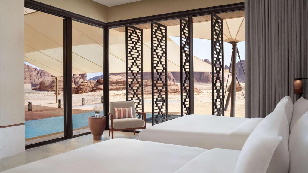 9 Best Hotels in Saudi Arabia: Unique Stays Across the Country 16 A modern hotel room with two beds, a chair, and floor-to-ceiling windows overlooks a private pool, sandy desert landscape, and distant rocky mountains—offering an experience among the best hotels in Saudi Arabia. Geometric screens provide shaded comfort outside.