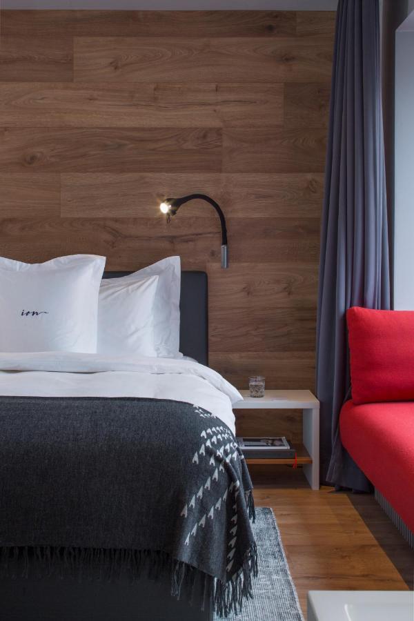 Where to Stay in Reykjavík: Top Guide 15 A modern hotel room—ideal for those seeking where to stay in Reykjavik—features a bed with white linens and a dark gray blanket, a wooden accent wall, reading lamp, nightstand with water, gray curtains, and a red couch.