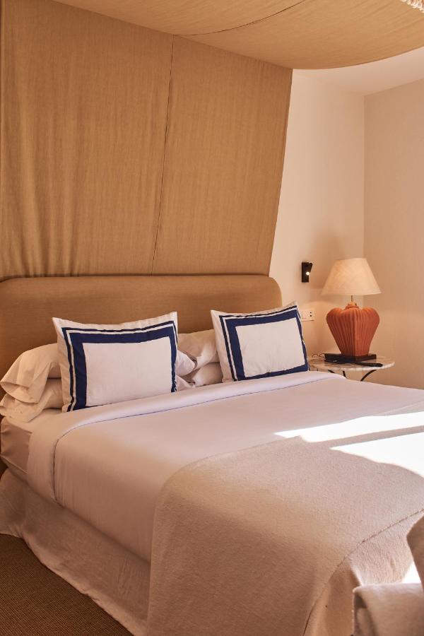 A neatly made bed with white linens and two pillows with blue borders sits against a tan upholstered headboard, capturing the inviting charm found in boutique hotels in Costa Brava. Sunlight streams onto the bed beside a lamp-lit table.