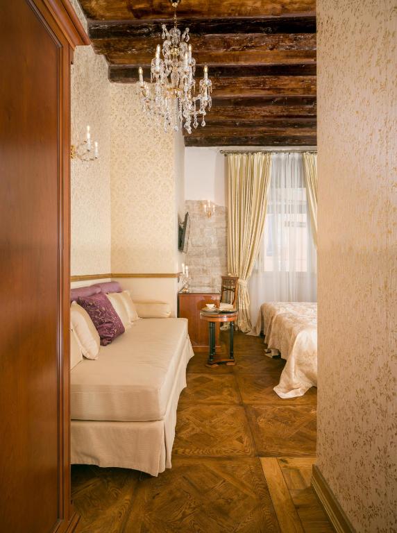 8 TOP Boutique Hotels in Split 7 A cozy, elegant room—perfect for those wondering where to stay in Split—with a cream-colored sofa, purple cushions, crystal chandelier, wooden beams, light floral wallpaper, and a bed with golden bedding near tall curtained windows.
