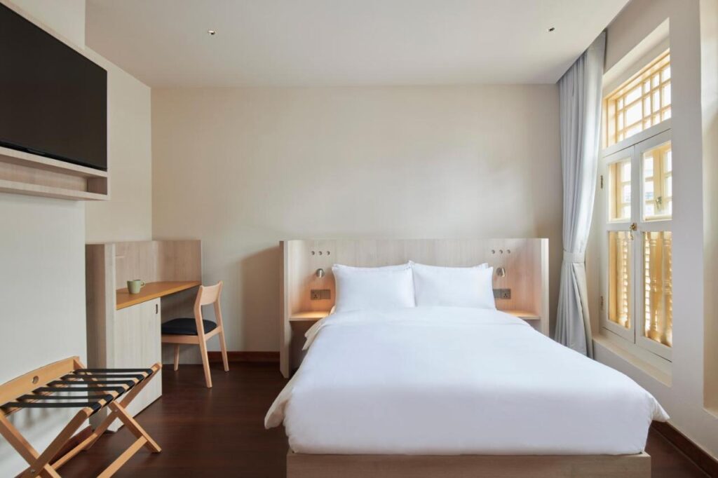 13 Most Unique Hotels in Singapore for an Incredible Holiday 8 A minimalist hotel room with a double bed, white bedding, a small desk and chair, a wall-mounted TV, and large windows with light curtains—perfect for guests seeking unique hotels in Singapore filled with natural light.