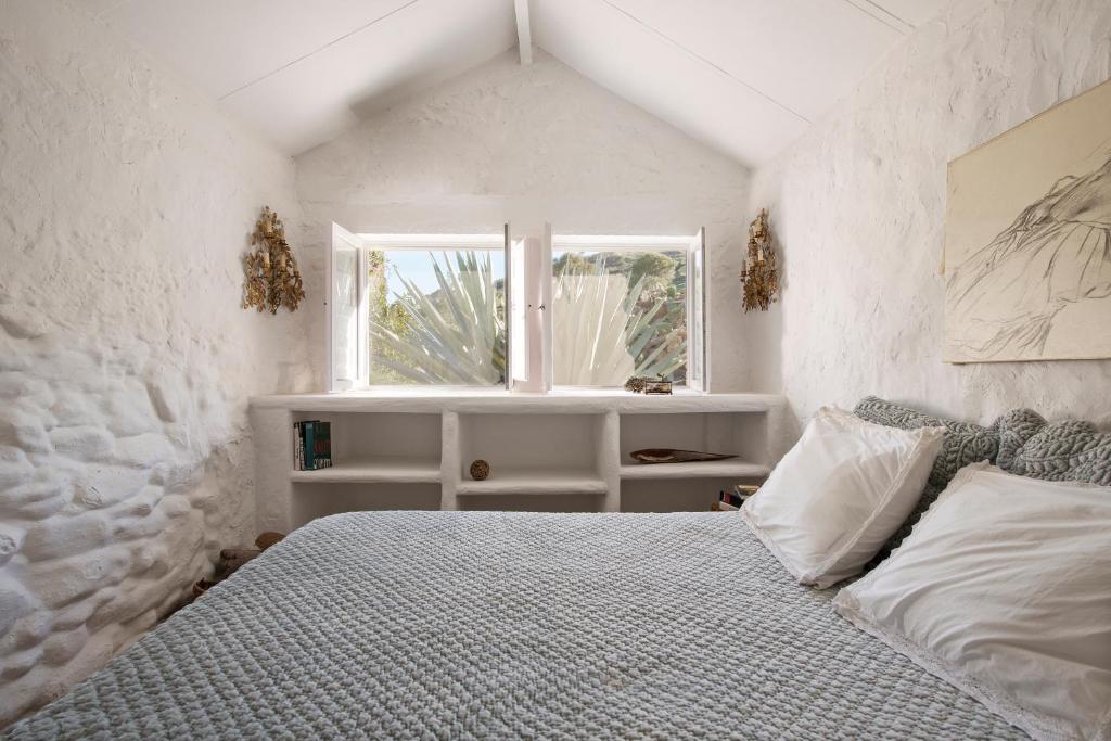 Adults-Only Hotels in Costa del Sol 39 Cozy bedroom with white stone walls, a bed with textured gray bedding, and open shelves—reminiscent of adult only hotels in Costa del Sol. A window lets in natural light and views of large agave plants. Dried plants and artwork decorate the space.
