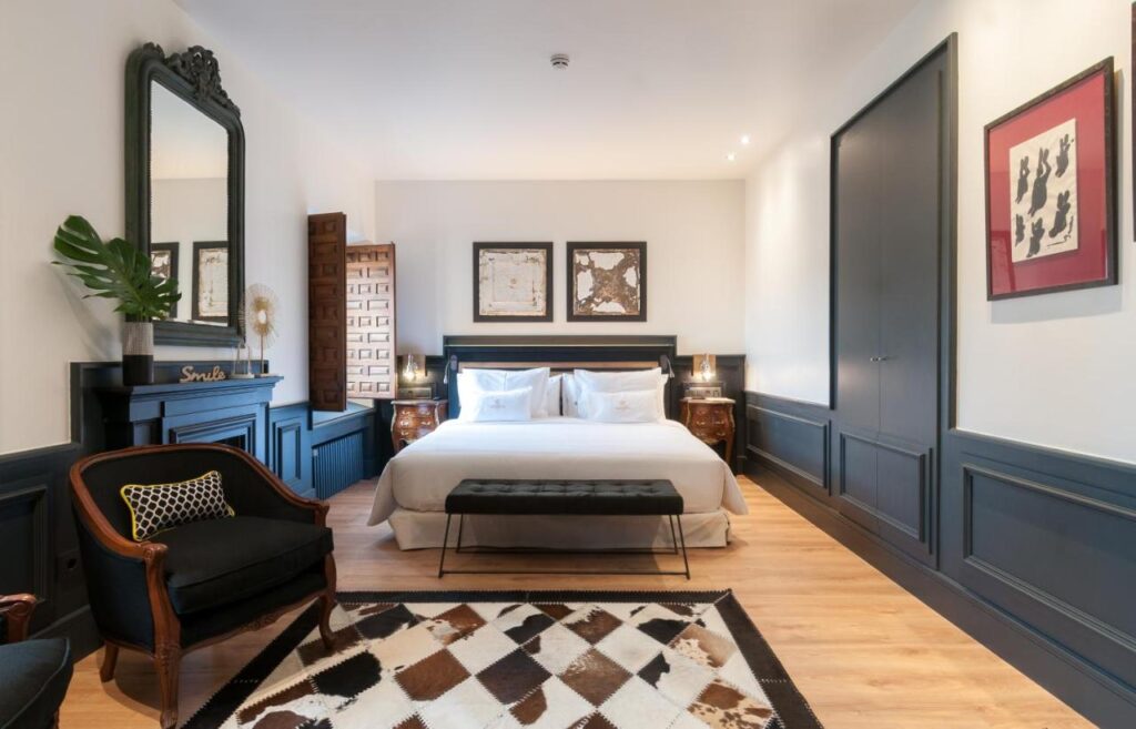 25 Weekend Trips from Madrid Max. 2h Away 98 A stylish bedroom inspired by weekend trips from Madrid, featuring a large bed with white bedding, dark blue paneled walls, wooden floor, a black bench, patterned rug, vintage armchair, decorative mirror, and framed artwork above the bed.